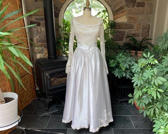 Könnte beinhalten: Ein weißes Satin-Hochzeitskleid mit langen Spitzenärmeln und einem Spitzenoberteil. Das Kleid hat einen vollen Rock und einen Spitzenbesatz am Saum.
