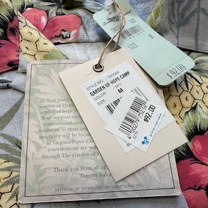 Może przedstawiać: Zbliżenie koszuli we wzory kwiatowe z dołączonymi metkami. Metki wskazują nazwę modelu "GARDEN OF HOPE CAMP" i kolor "SKY". Koszula jest w rozmiarze M.