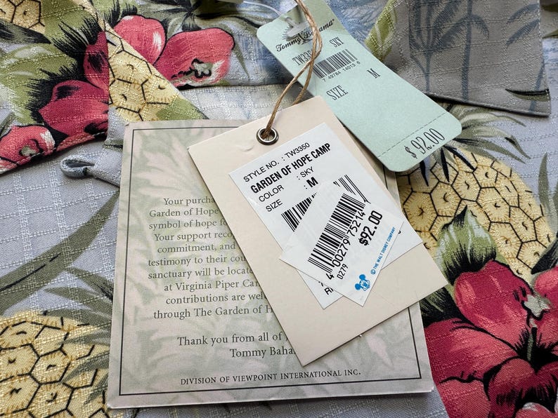 Może przedstawiać: Zbliżenie koszuli we wzory kwiatowe z dołączonymi metkami. Metki wskazują nazwę modelu "GARDEN OF HOPE CAMP" i kolor "SKY". Koszula jest w rozmiarze M.