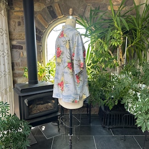 Może przedstawiać: Jasnoniebieska koszula hawajska z kwiatowym wzorem z czerwonych i ż&oacute;łtych kwiat&oacute;w oraz zielonych liści. Koszula jest prezentowana na manekinie. Koszula ma kr&oacute;tkie rękawy i zapinany na guziki prz&oacute;d.