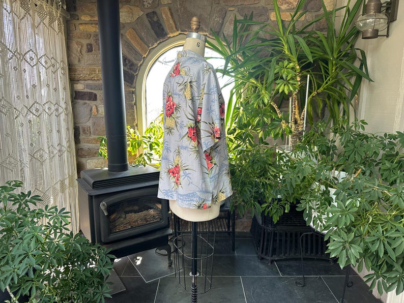 Może przedstawiać: Jasnoniebieska koszula hawajska z kwiatowym wzorem z czerwonych i ż&oacute;łtych kwiat&oacute;w oraz zielonych liści. Koszula jest prezentowana na manekinie. Koszula ma kr&oacute;tkie rękawy i zapinany na guziki prz&oacute;d.