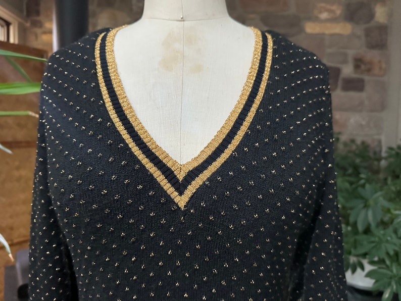 Pu&ograve; includere: Maglione nero a maglia con scollo a V e dettagli dorati. Il maglione ha un bordo dorato intorno allo scollo e piccoli punti dorati su tutto il tessuto. Il maglione &egrave; esposto su un manichino.