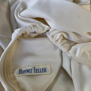 Peut inclure: Gros plan sur un v&ecirc;tement cr&egrave;me avec une fermeture &eacute;clair et une &eacute;tiquette qui indique "BONWIT TELLER" en texte bleu. Le tissu semble &ecirc;tre un mat&eacute;riau lisse et tiss&eacute;. Le v&ecirc;tement est partiellement ouvert, r&eacute;v&eacute;lant la doublure int&eacute;rieure.