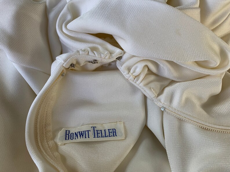 Peut inclure: Gros plan sur un v&ecirc;tement cr&egrave;me avec une fermeture &eacute;clair et une &eacute;tiquette qui indique "BONWIT TELLER" en texte bleu. Le tissu semble &ecirc;tre un mat&eacute;riau lisse et tiss&eacute;. Le v&ecirc;tement est partiellement ouvert, r&eacute;v&eacute;lant la doublure int&eacute;rieure.