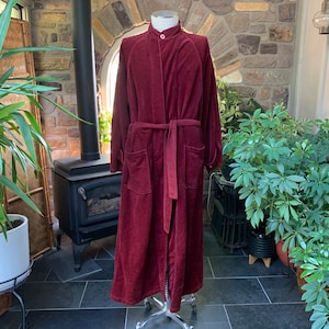 May include: A long-sleeved, floor-length burgundy robe made of a textured fabric. The garment features a high mandarin collar with a single white button closure at the neck, a matching tie belt at the waist, and two large patch pockets on the front.