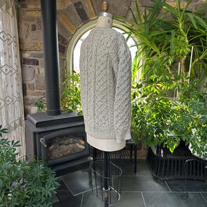 K&ouml;nnte beinhalten: Hellgrauer Zopfstrickpullover auf einer Schaufensterpuppe. Der Pullover hat ein strukturiertes Muster und lange &Auml;rmel. Die Schaufensterpuppe steht auf einem schwarzen Metallst&auml;nder. Der Hintergrund umfasst einen Kamin, Pflanzen und ein Fenster.