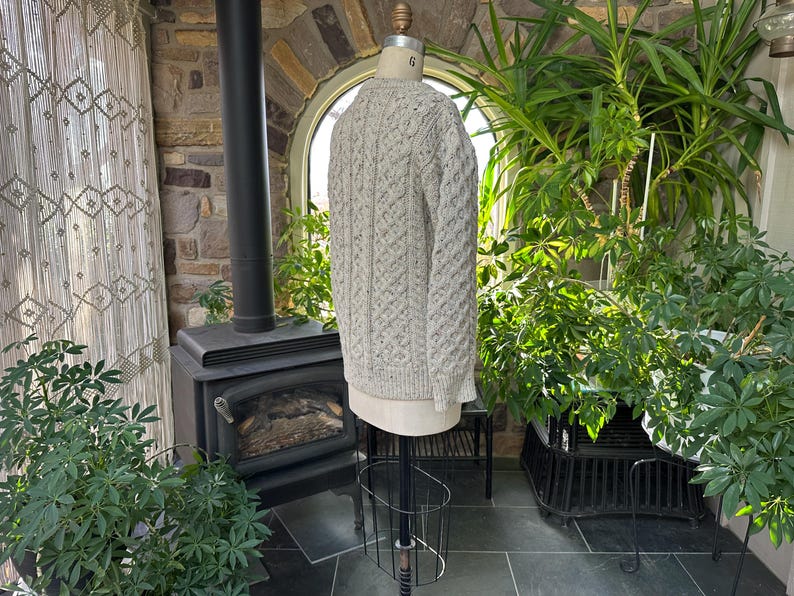 K&ouml;nnte beinhalten: Hellgrauer Zopfstrickpullover auf einer Schaufensterpuppe. Der Pullover hat ein strukturiertes Muster und lange &Auml;rmel. Die Schaufensterpuppe steht auf einem schwarzen Metallst&auml;nder. Der Hintergrund umfasst einen Kamin, Pflanzen und ein Fenster.