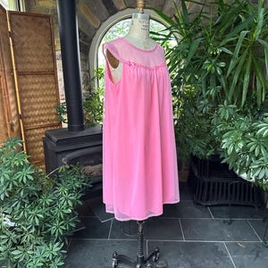 Puede incluir: Un camis&oacute;n rosa vintage en un maniqu&iacute;. El camis&oacute;n es hasta la rodilla con una superposici&oacute;n transparente y ribete de encaje. El maniqu&iacute; est&aacute; sobre un soporte con ruedas. El fondo incluye una chimenea, plantas y una pantalla de bamb&uacute;.