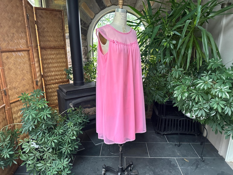 Puede incluir: Un camis&oacute;n rosa vintage en un maniqu&iacute;. El camis&oacute;n es hasta la rodilla con una superposici&oacute;n transparente y ribete de encaje. El maniqu&iacute; est&aacute; sobre un soporte con ruedas. El fondo incluye una chimenea, plantas y una pantalla de bamb&uacute;.