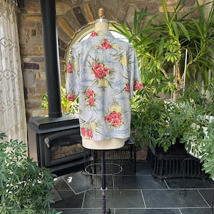 Może przedstawiać: Jasnoniebieska koszula hawajska z kwiatowym wzorem czerwonych kwiat&oacute;w hibiskusa i ż&oacute;łtych ananas&oacute;w. Koszula z kr&oacute;tkim rękawem jest prezentowana na manekinie, prezentując sw&oacute;j design. Koszula jest wykonana z lekkiego materiału.