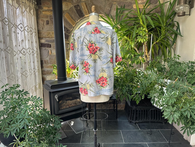 Może przedstawiać: Jasnoniebieska koszula hawajska z kwiatowym wzorem czerwonych kwiat&oacute;w hibiskusa i ż&oacute;łtych ananas&oacute;w. Koszula z kr&oacute;tkim rękawem jest prezentowana na manekinie, prezentując sw&oacute;j design. Koszula jest wykonana z lekkiego materiału.