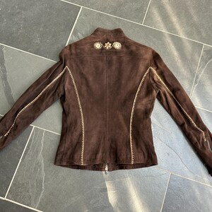 May include: A brown suede jacket with decorative stitching and embellishments on the shoulders. The jacket features long sleeves with zippers at the cuffs and a full-length zipper closure. The stitching is a contrasting light color.