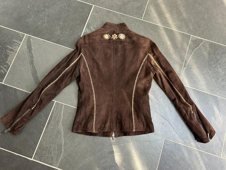 May include: A brown suede jacket with decorative stitching and embellishments on the shoulders. The jacket features long sleeves with zippers at the cuffs and a full-length zipper closure. The stitching is a contrasting light color.