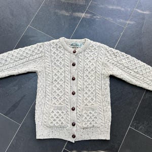 K&ouml;nnte beinhalten: Hellgraue Strickjacke mit Knopfleiste und Zopfmuster. Der Pullover hat zwei Vordertaschen und braune Kn&ouml;pfe. Die langen &Auml;rmel und der Stil sind klassisch und warm.