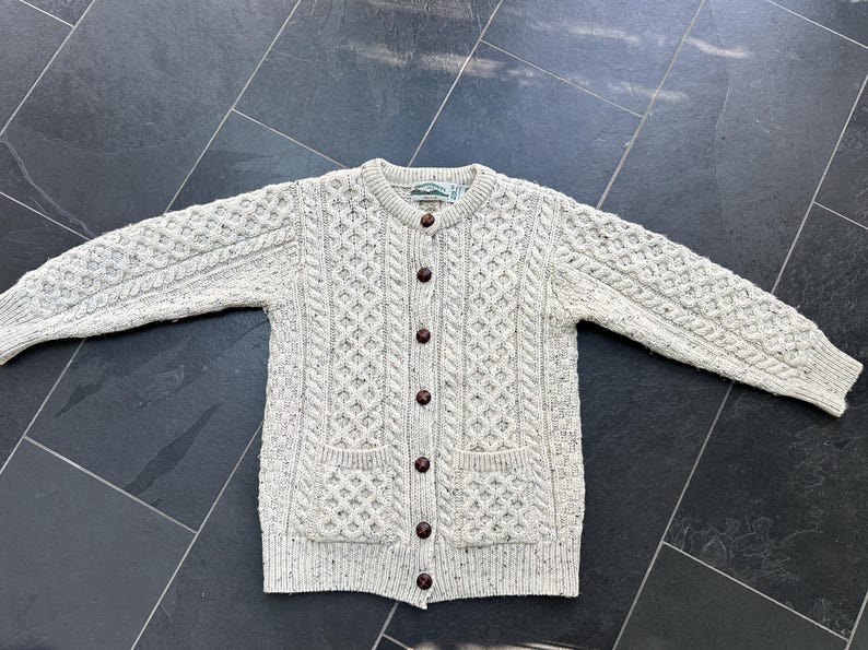 K&ouml;nnte beinhalten: Hellgraue Strickjacke mit Knopfleiste und Zopfmuster. Der Pullover hat zwei Vordertaschen und braune Kn&ouml;pfe. Die langen &Auml;rmel und der Stil sind klassisch und warm.
