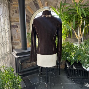 May include: A dark brown suede jacket with long sleeves and decorative stitching. The jacket features floral embroidery on the collar and is displayed on a mannequin. The back view showcases the tailored design and detailed craftsmanship.