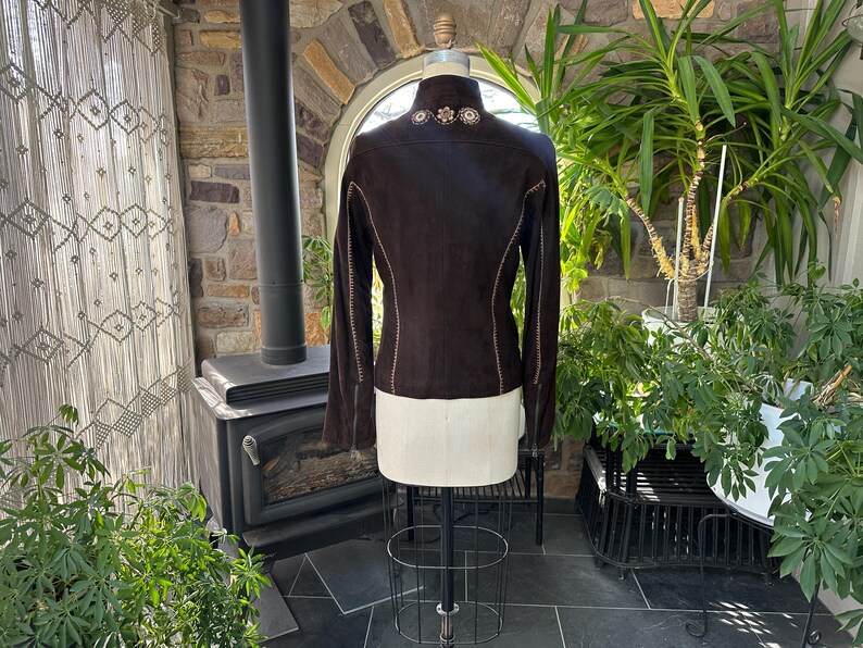 May include: A dark brown suede jacket with long sleeves and decorative stitching. The jacket features floral embroidery on the collar and is displayed on a mannequin. The back view showcases the tailored design and detailed craftsmanship.