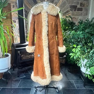 Puede incluir: Un abrigo largo de color tostado con un grueso ribete de piel de oveja color crema a lo largo del cuello, la abertura frontal, los puños y el dobladillo. El abrigo incluye detalles de costuras decorativas a lo largo de las uniones y se muestra en un maniquí.