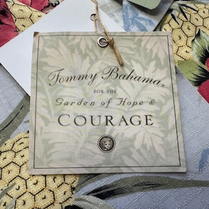 Może przedstawiać: Kwadratowa etykieta Tommy Bahama z czarnym napisem "Tommy Bahama. For the Garden of Hope & Courage". Etykieta jest kremowa z wzorem liści i brązowym sznurkiem. Tło przedstawia tkaninę kwiatową.