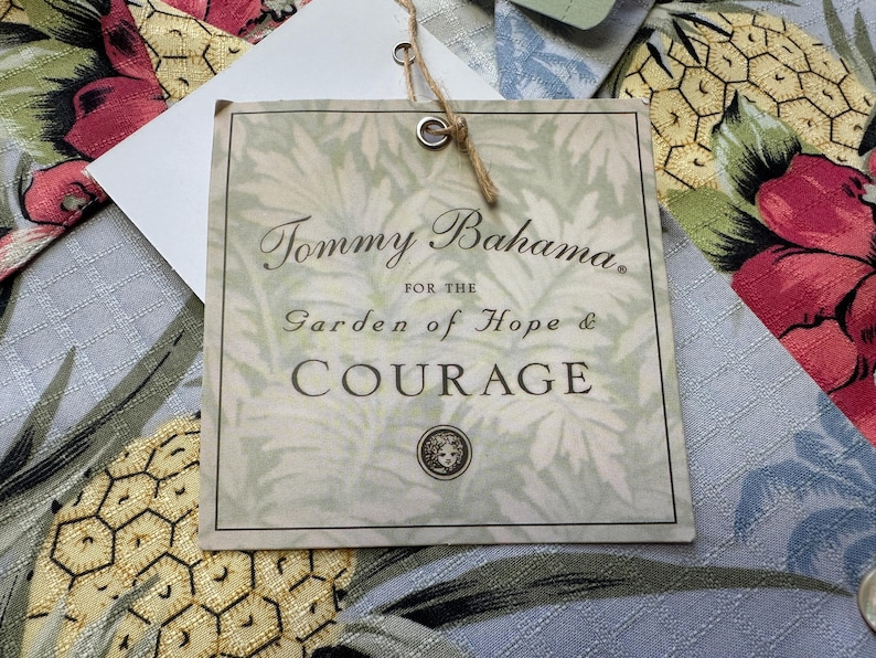 Może przedstawiać: Kwadratowa etykieta Tommy Bahama z czarnym napisem "Tommy Bahama. For the Garden of Hope & Courage". Etykieta jest kremowa z wzorem liści i brązowym sznurkiem. Tło przedstawia tkaninę kwiatową.