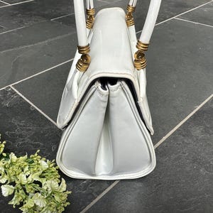 May include: A white handbag with a glossy finish, featuring a structured gusseted side profile and two white handles attached with gold-toned metal hardware. The bag has a flap closure and a smooth, clean exterior surface with visible stitching.