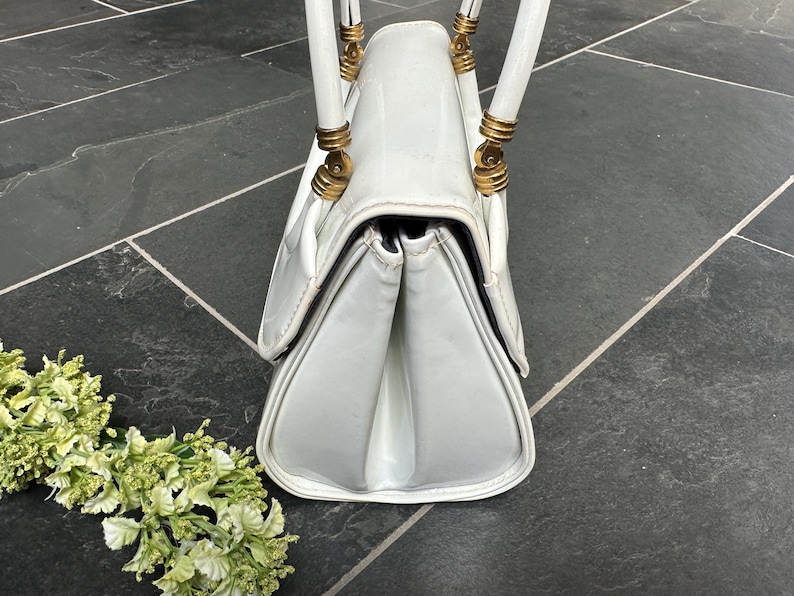 May include: A white handbag with a glossy finish, featuring a structured gusseted side profile and two white handles attached with gold-toned metal hardware. The bag has a flap closure and a smooth, clean exterior surface with visible stitching.