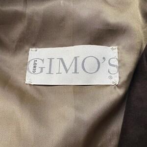 May include: Close-up of a label inside a garment. The label is rectangular, white, and features the brand name "GIMO'S" in gray lettering. The label is sewn onto a tan fabric, with a dark brown fabric visible on the right side.