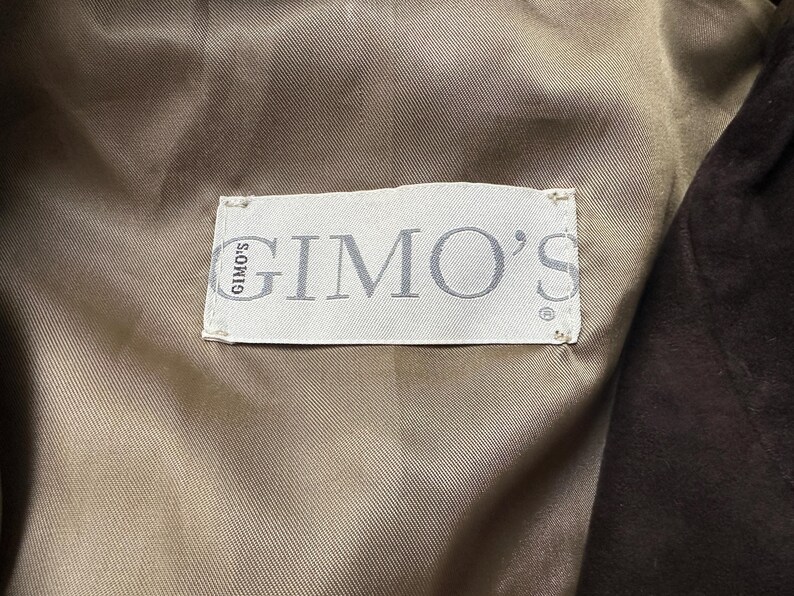 May include: Close-up of a label inside a garment. The label is rectangular, white, and features the brand name "GIMO'S" in gray lettering. The label is sewn onto a tan fabric, with a dark brown fabric visible on the right side.