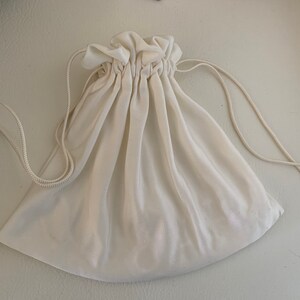 Peut inclure: Pochette &agrave; cordon de serrage blanc cass&eacute;. Le sac est fait d'un tissu doux et semi-transparent. Le haut du sac est fronc&eacute; avec une fermeture &agrave; cordon. Le cordon est une fine corde blanc cass&eacute;. Le sac est vide et pos&eacute; sur une surface blanche.