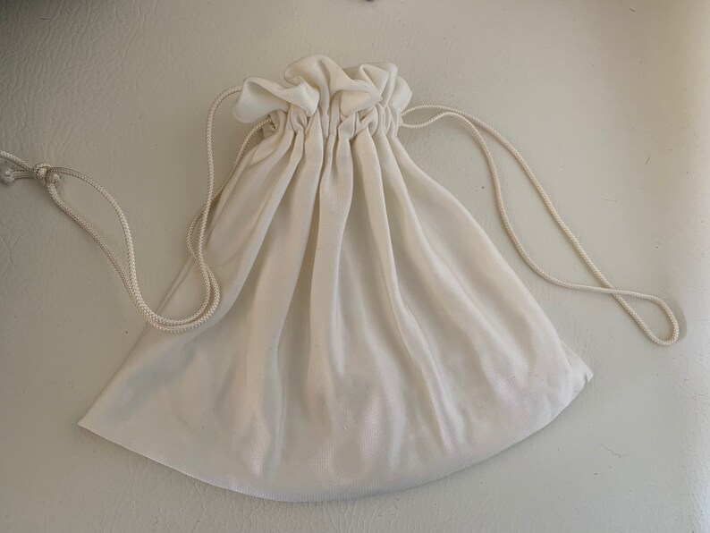 Peut inclure: Pochette &agrave; cordon de serrage blanc cass&eacute;. Le sac est fait d'un tissu doux et semi-transparent. Le haut du sac est fronc&eacute; avec une fermeture &agrave; cordon. Le cordon est une fine corde blanc cass&eacute;. Le sac est vide et pos&eacute; sur une surface blanche.