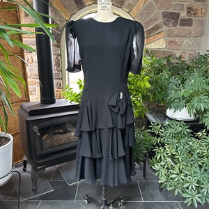 May include: A black tiered dress with sheer sleeves and a bow detail at the waist. The dress is on a mannequin in a room with plants and a fireplace.