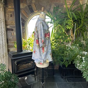 Może przedstawiać: Jasnoniebieska koszula w kwiatowy wz&oacute;r z czerwonymi i ż&oacute;łtymi kwiatami. Koszula jest prezentowana na manekinie. Ma kr&oacute;tkie rękawy i kołnierzyk. Tło obejmuje kominek, rośliny i okno. Rozmiar 6.