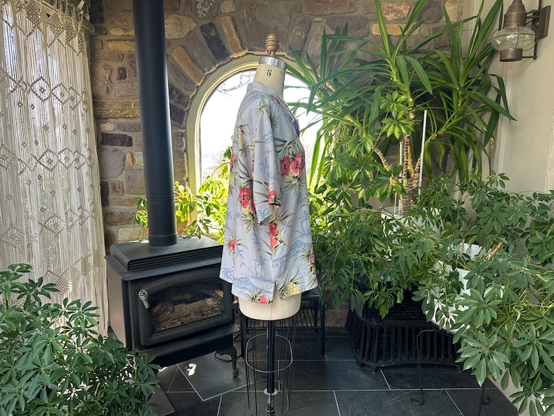 Może przedstawiać: Jasnoniebieska koszula w kwiatowy wz&oacute;r z czerwonymi i ż&oacute;łtymi kwiatami. Koszula jest prezentowana na manekinie. Ma kr&oacute;tkie rękawy i kołnierzyk. Tło obejmuje kominek, rośliny i okno. Rozmiar 6.