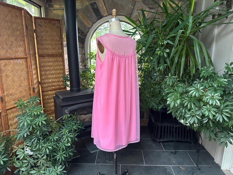 Puede incluir: Un camis&oacute;n rosa vintage en un maniqu&iacute;. El camis&oacute;n sin mangas tiene un canes&uacute; transparente con adornos decorativos y una falda fluida hasta la rodilla. La tela parece ligera y el estilo es cl&aacute;sico.