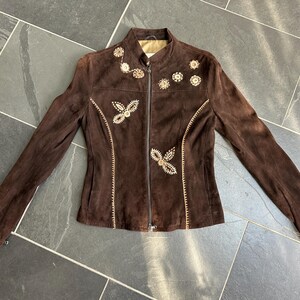 May include: A brown suede jacket with a zip-up front and long sleeves. The jacket features embroidered floral and geometric designs in a lighter brown color. The jacket is laid flat on a dark gray tiled surface.