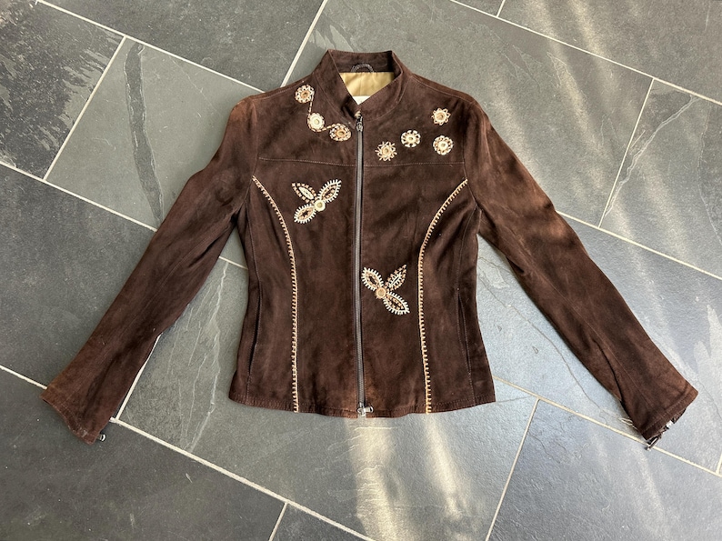 May include: A brown suede jacket with a zip-up front and long sleeves. The jacket features embroidered floral and geometric designs in a lighter brown color. The jacket is laid flat on a dark gray tiled surface.