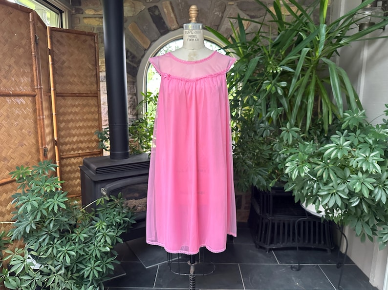 Puede incluir: Un camis&oacute;n rosa vintage en un maniqu&iacute;. El camis&oacute;n tiene un canes&uacute; transparente y mangas cortas con un ribete con volantes. El vestido largo y fluido est&aacute; hecho de una tela ligera. El maniqu&iacute; est&aacute; sobre un soporte de metal, con plantas y una chimenea en el fondo.