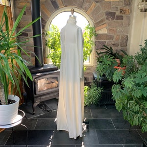 Peut inclure: Une longue robe ivoire &agrave; col montant et manches longues. La robe est pr&eacute;sent&eacute;e sur un mannequin dans une pi&egrave;ce ensoleill&eacute;e avec un mur en pierre et diverses plantes en pot. La robe a une silhouette fluide.