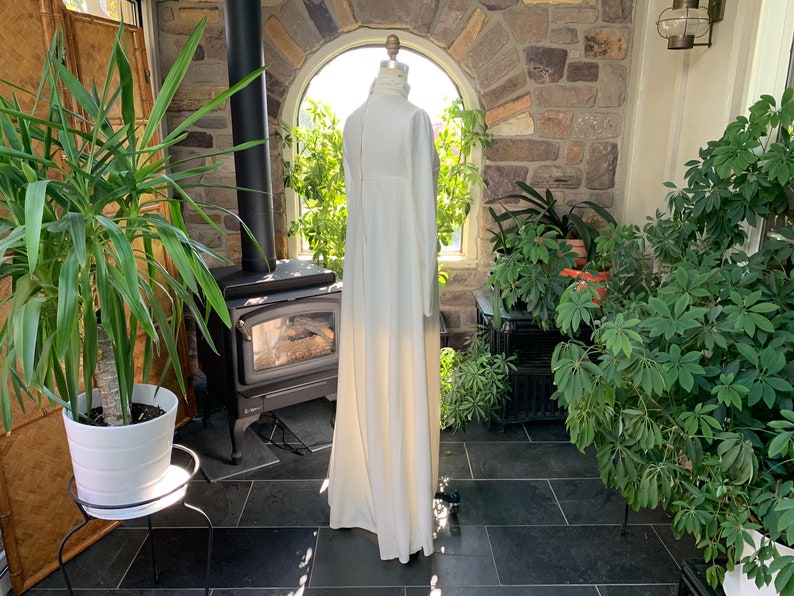 Peut inclure: Une longue robe ivoire &agrave; col montant et manches longues. La robe est pr&eacute;sent&eacute;e sur un mannequin dans une pi&egrave;ce ensoleill&eacute;e avec un mur en pierre et diverses plantes en pot. La robe a une silhouette fluide.