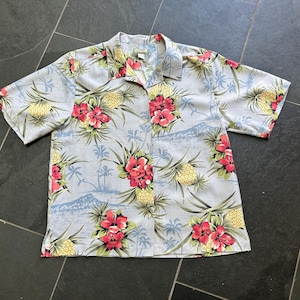 Może przedstawiać: Jasnoniebieska koszula z kr&oacute;tkim rękawem z tropikalnym nadrukiem kwiatowym. Koszula ma wz&oacute;r czerwonych kwiat&oacute;w hibiskusa, ż&oacute;łtych ananas&oacute;w i palm. Ma zapięcie na guziki i kołnierzyk.