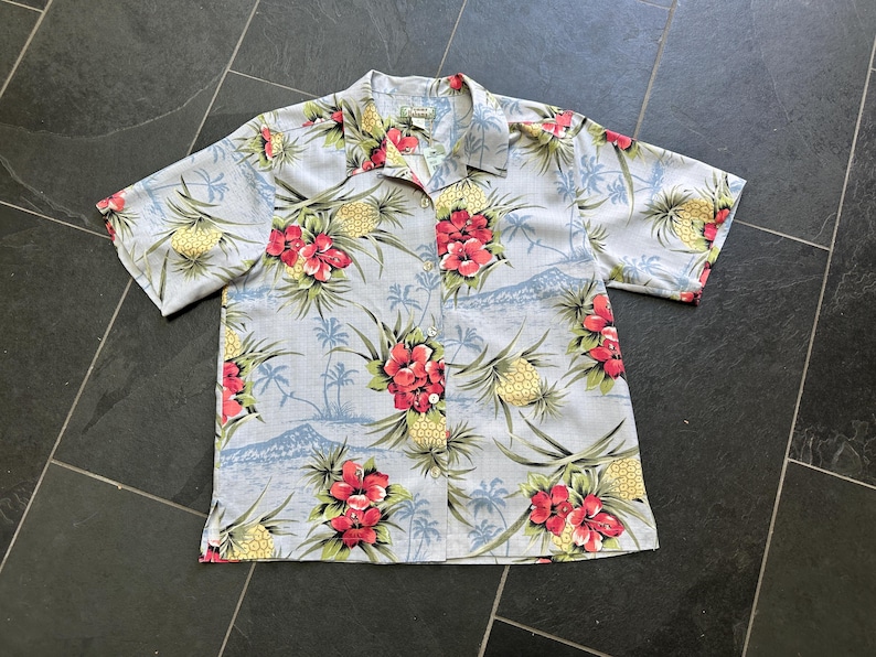 Może przedstawiać: Jasnoniebieska koszula z kr&oacute;tkim rękawem z tropikalnym nadrukiem kwiatowym. Koszula ma wz&oacute;r czerwonych kwiat&oacute;w hibiskusa, ż&oacute;łtych ananas&oacute;w i palm. Ma zapięcie na guziki i kołnierzyk.