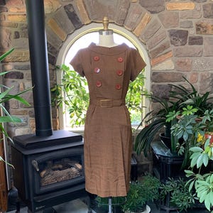 May include: A brown dress with a short sleeve, a belt, and a double row of buttons. The dress is on a mannequin in front of a stone wall.
