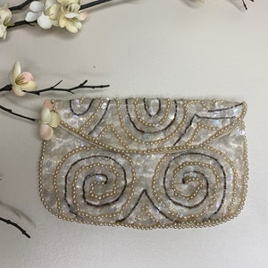 May include: A white, sequined clutch with a flap closure. The clutch features a swirling design created with black beads and small, white pearls. The bag is decorated with small, white flowers on the side. A stylish accessory for special occasions.