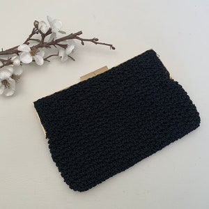 May include: A black, crocheted clutch with a gold-toned clasp. The rectangular bag has a textured surface and is set against a white background. A sprig of white flowers is in the upper left corner.