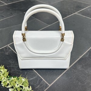 May include: A glossy white handbag with a structured rectangular body and a flap closure. The bag features a single rounded top handle attached with metallic gold-toned hardware rings. The surface has a smooth, reflective finish throughout.