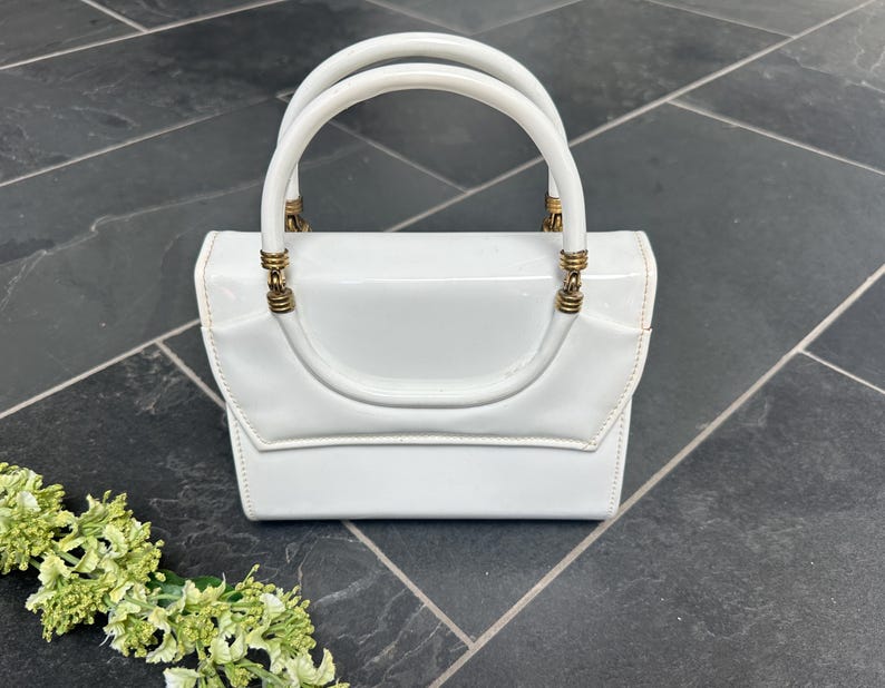 May include: A glossy white handbag with a structured rectangular body and a flap closure. The bag features a single rounded top handle attached with metallic gold-toned hardware rings. The surface has a smooth, reflective finish throughout.