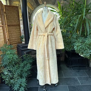 Pode incluir: Um roupão longo, amarelo claro, com gola de xale e cinto a condizer. O roupão é exibido num manequim. O roupão é feito de um material macio e felpudo. O roupão mede aproximadamente 140 cm de comprimento.