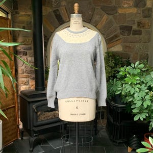 May include: A light gray sweater with a cream-colored yoke, displayed on a mannequin. The yoke is adorned with small, round embellishments. The sweater has long sleeves and a classic crew neck. The mannequin is on a metal stand, and the background features a brick wall and greenery.