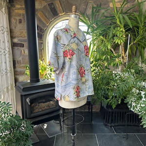 Może przedstawiać: Jasnoniebieska koszula hawajska z tropikalnym nadrukiem kwiatowym, w tym czerwone kwiaty i ananasy. Koszula jest prezentowana na manekinie, prezentując kr&oacute;tkie rękawy i styl zapinany na guziki. Tło obejmuje rośliny i kamienną ścianę.