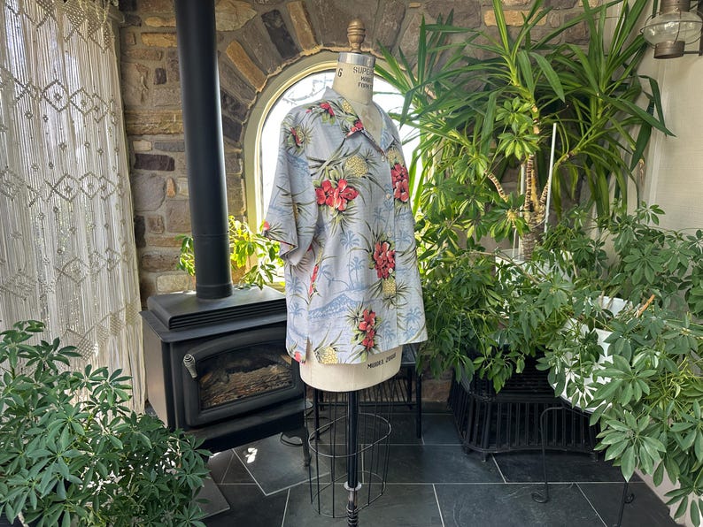 Może przedstawiać: Jasnoniebieska koszula hawajska z tropikalnym nadrukiem kwiatowym, w tym czerwone kwiaty i ananasy. Koszula jest prezentowana na manekinie, prezentując kr&oacute;tkie rękawy i styl zapinany na guziki. Tło obejmuje rośliny i kamienną ścianę.